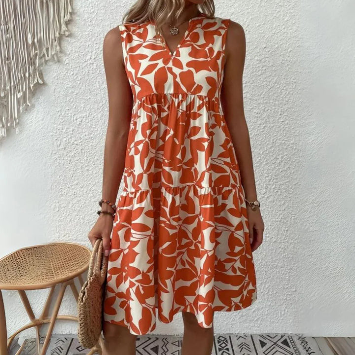 A woman wears a sleeveless dress in an orange and white abstract leaf pattern, perfect for a summer wardrobe. She holds a woven handbag and stands against a white textured wall near a wicker stool and macrame wall hanging.
