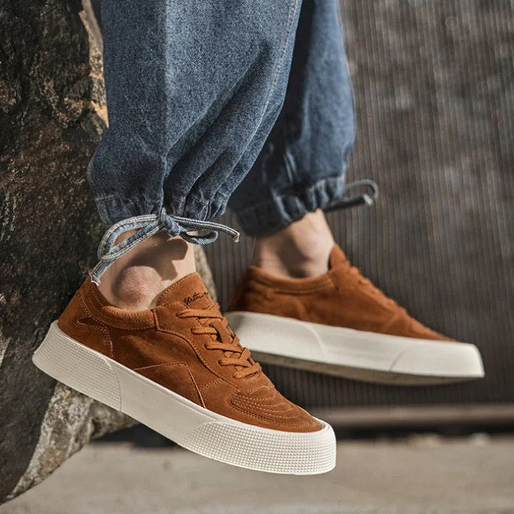 Person wearing blue cuffed jeans and brown suede lace-up sneakers with white soles, featuring comfortable cushioning, stands outside with one foot resting against a rock or concrete surface.