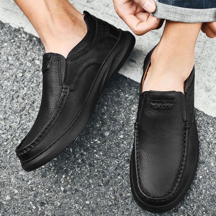 A person wears black slip-on men's cowhide leather shoes with visible stitching and a soft sole, standing on a textured gray asphalt surface while adjusting one pant leg.