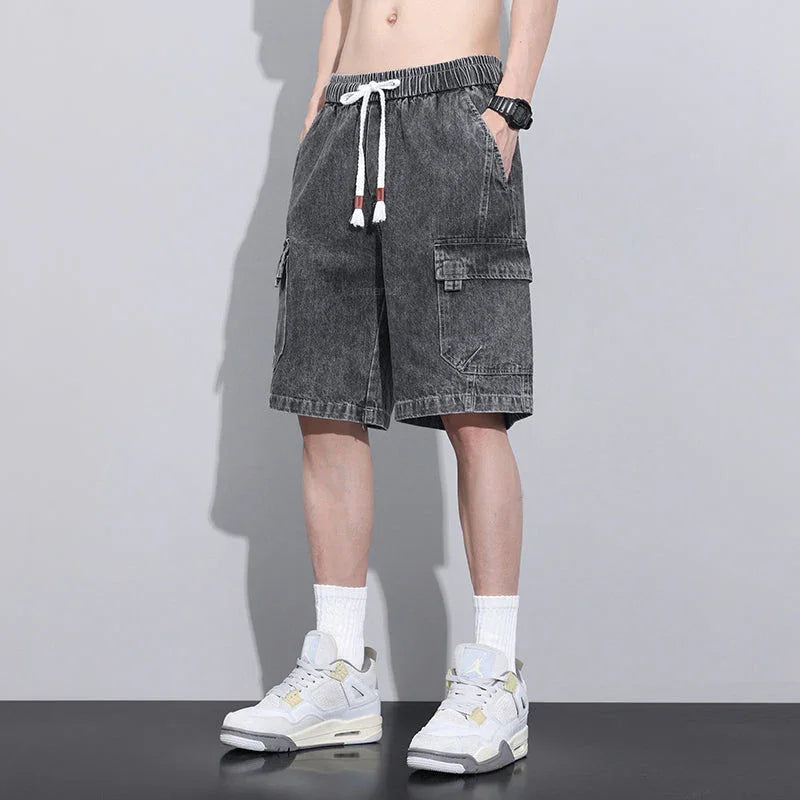 A person wearing black washed mens denim shorts with an elastic waist, white socks, white sneakers, a black watch, and a white drawstring, standing against a plain light gray background.