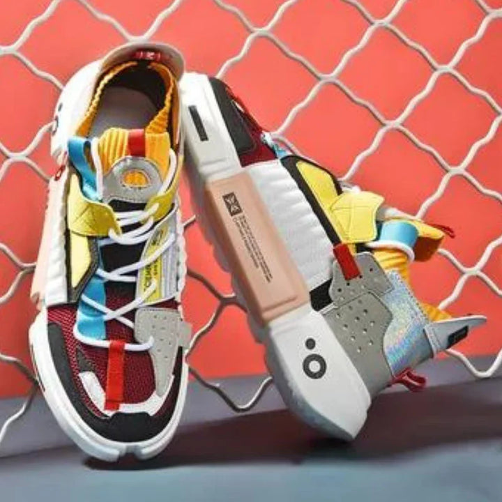 A pair of colorful, modern Extreme VS7 Sneakers with a mix of red, yellow, blue, black, and gray panels, white soles, and unique design elements—perfect lightweight breathable sneakers—displayed against a wavy metal fence and coral-red background.