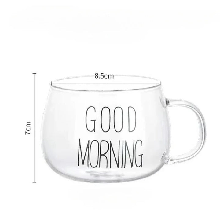 A clear glass mug with an ergonomic handle and GOOD MORNING written in black letters. Made from heat-resistant glass, the mug measures 8.5 cm in diameter and 7 cm in height, with a handle on the right side.
