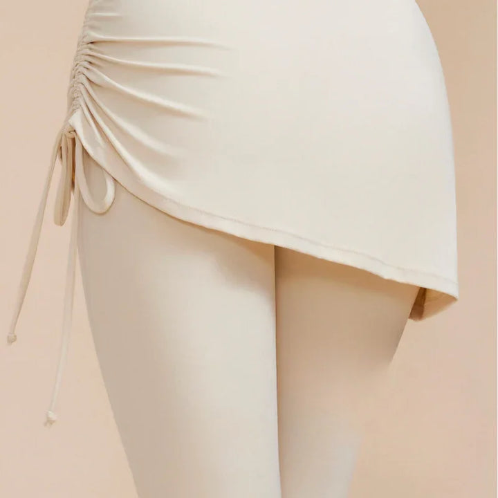 A close-up of a person wearing cream-colored, high-waisted leggings with skirt in a ruched, asymmetrical style. The skirt features a drawstring detail on one side, perfect for elevating your autumn wardrobe against a neutral beige background.