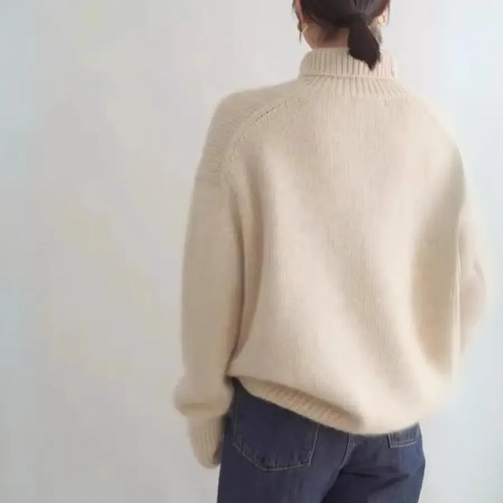 A person with dark hair tied back is wearing a loose, oversized fit beige turtleneck sweater and dark jeans, standing against a plain light-colored background with their back to the camera.