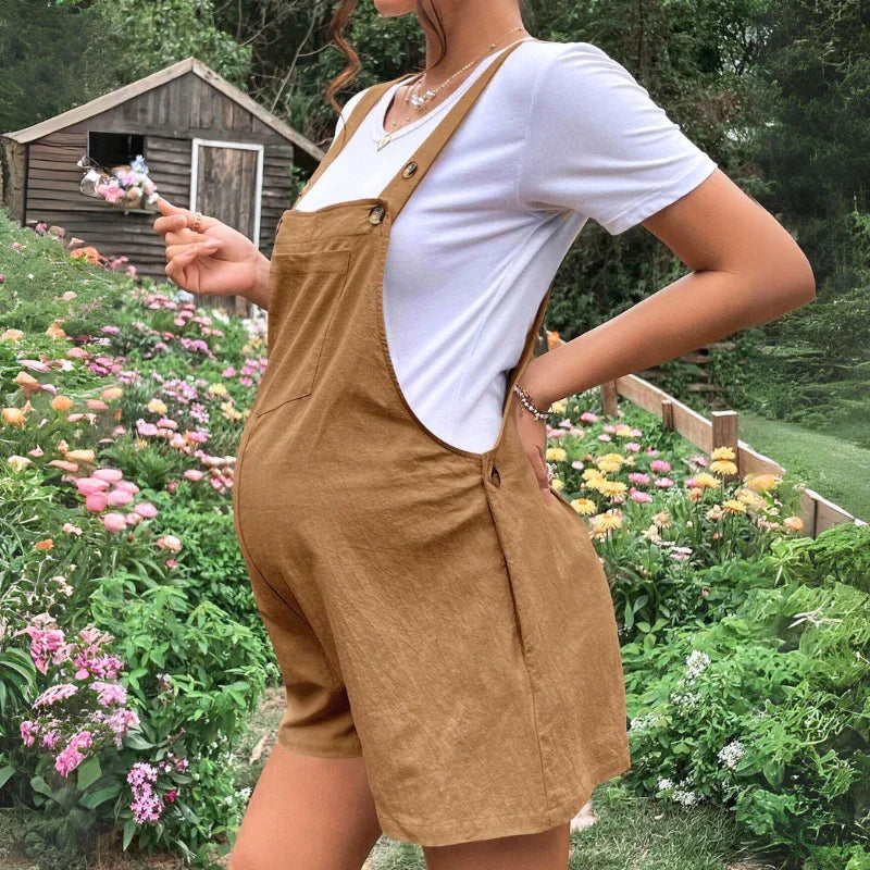 A pregnant woman in comfortable maternity clothes stands in a garden full of colorful flowers, holding a lollipop, with a wooden shed in the background.