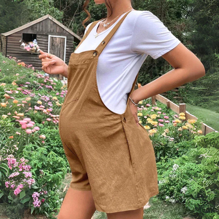 A pregnant woman in comfortable maternity clothes stands in a garden full of colorful flowers, holding a lollipop, with a wooden shed in the background.