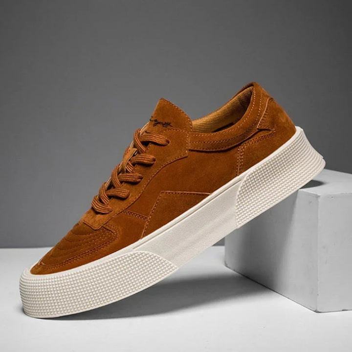 A brown suede lace-up sneaker with matching laces and a thick white textured rubber sole is displayed against a gray background, propped up on a white rectangular block for comfortable cushioning.
