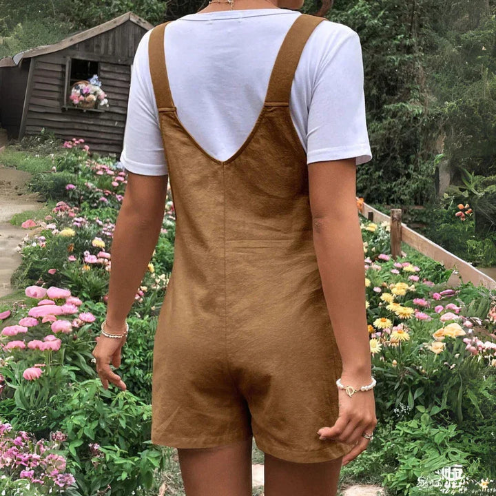 A person stands in a garden full of colorful flowers, wearing comfortable maternity clothes—a white short-sleeve shirt under brown maternity overalls. The scene includes a wooden shed and lush greenery in the background. The person is seen from the back.