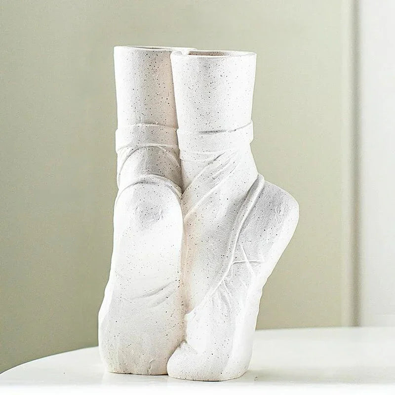 A white ceramic vase resembling two ballet slippers, posed en pointe, sits on a smooth white surface against a light, blurred background—perfect for contemporary chic décor.