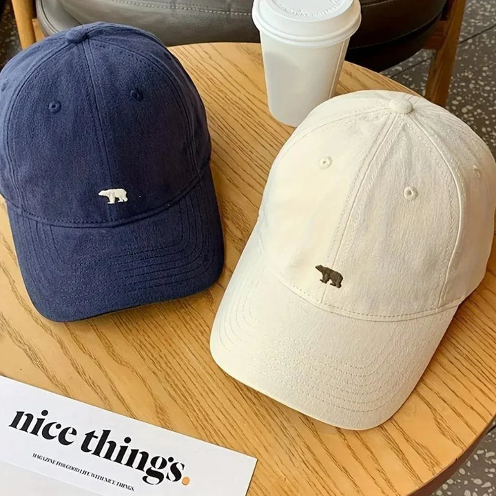 Two premium cotton baseball caps, one navy blue and one cream, each with a small bear logo on the front, rest on a wooden table beside a white coffee cup and a magazine titled nice things—perfect for stylish sun protection.