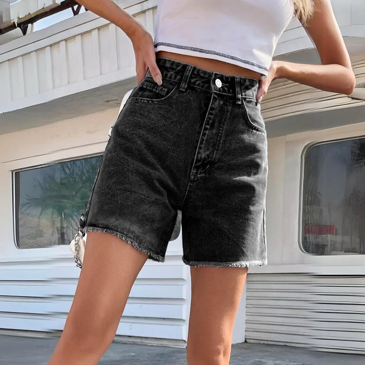 A person wearing a white crop top and high-waist black wash denim streetwear shorts stands outside in front of a white modern building. Their face is not visible in the image.
