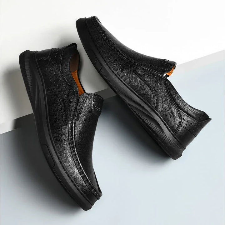 A pair of men's cowhide leather slip-on shoes with textured uppers and thick soft soles, positioned diagonally on a white and light blue background.