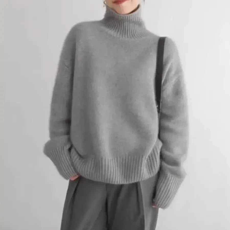 A person wearing a loose-fitting, thick knit gray turtleneck sweater and dark gray pants stands against a plain white background.