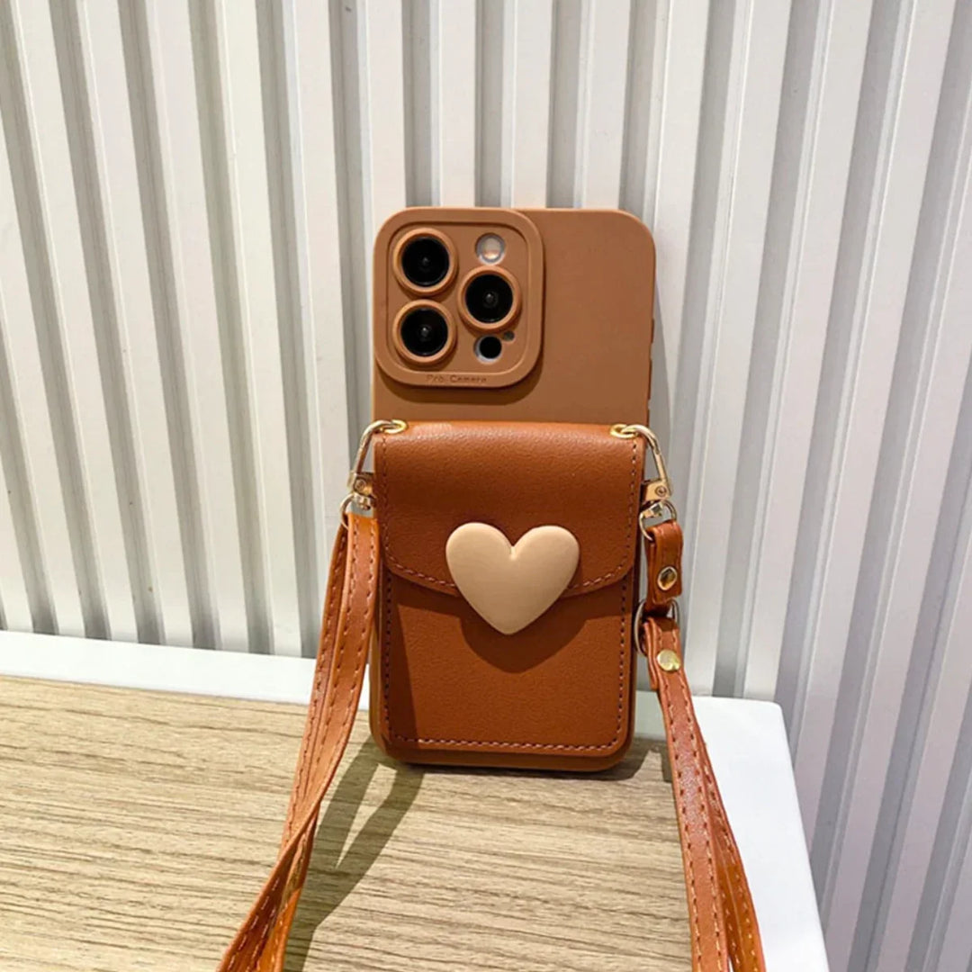 A chic phone case with a crossbody strap and a small heart-shaped pocket sits upright on a wooden surface, its brown design complemented by a beige heart, against a white, ridged wall.