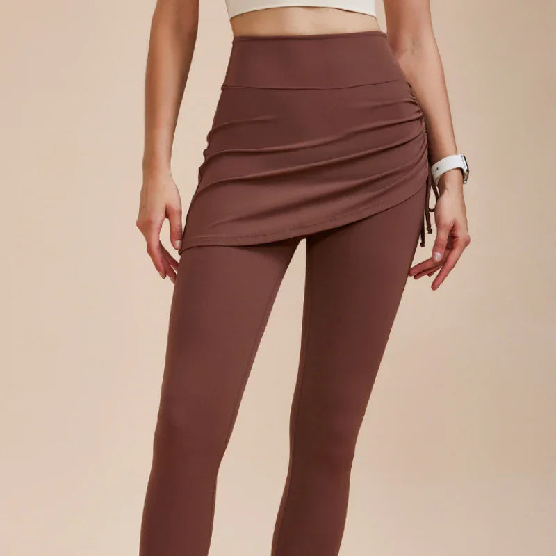 A person wears high-waisted leggings with skirt in brown, paired with a white crop top, standing against a plain beige background. Their face is not visible—a chic autumn wardrobe staple.