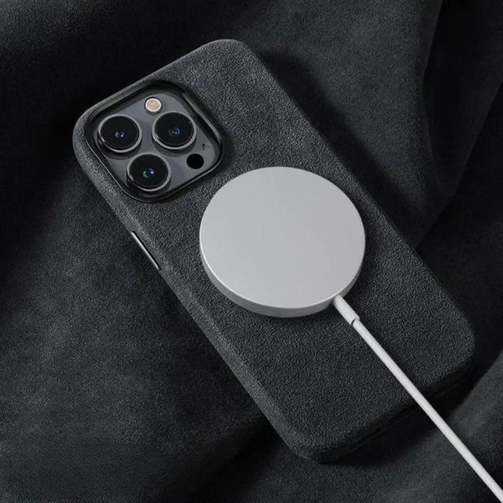 A smartphone with a dark, suede finish case rests on a soft, dark surface while a round, magnetic wireless charger is attached to its back.