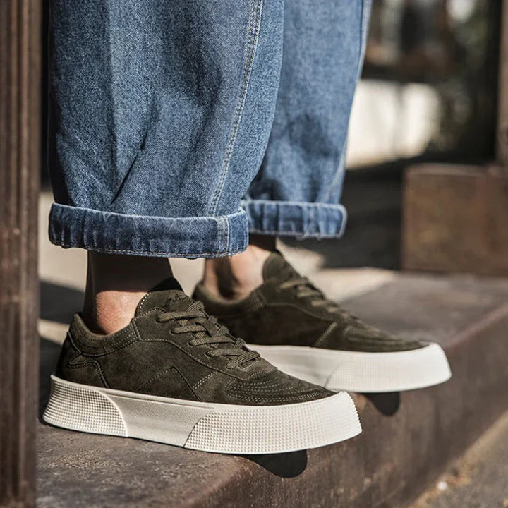 A person wearing cuffed blue jeans and olive green lace-up sneakers with comfortable cushioning and thick white soles stands outdoors on a concrete surface.
