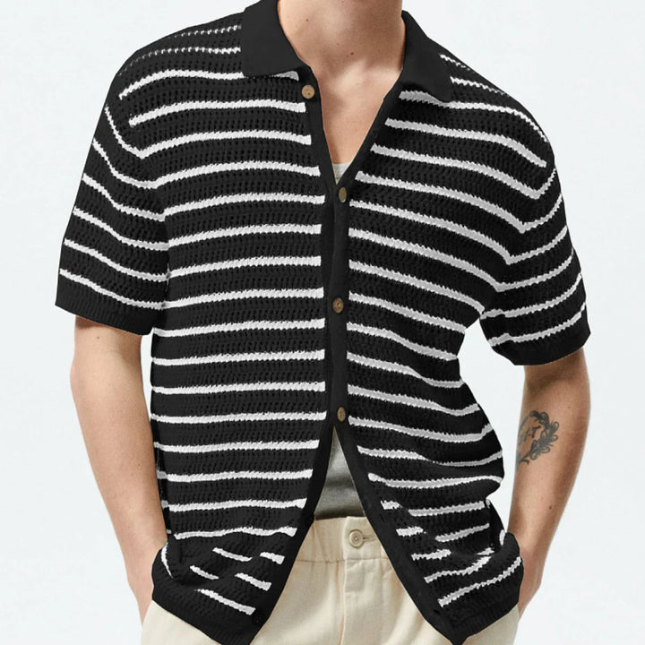 A person wearing a black retro knit polo with horizontal white stripes, beige pants, and a visible tattoo on their left forearm. The shirt features an open-knit design and is unbuttoned, revealing a gray undershirt.