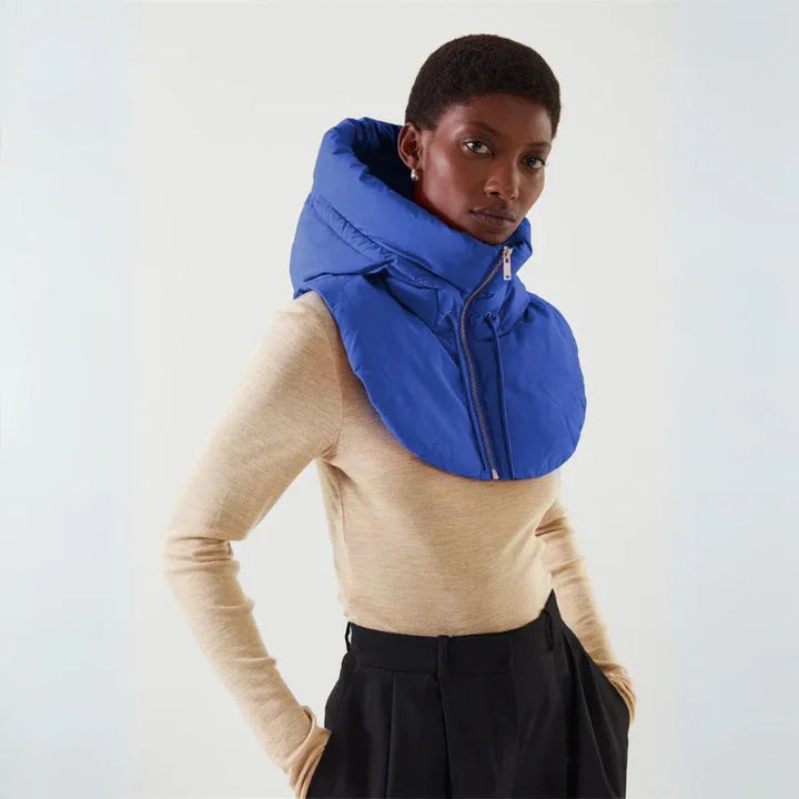 A person wearing a beige long-sleeve top, black pants, and a large blue puffer scarf made of durable material with a high collar and zipper, standing against a plain light background.