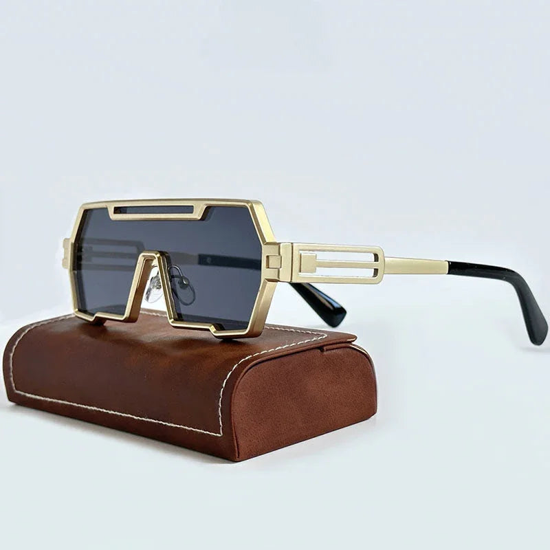Geometric gold-framed steampunk sunglasses with dark lenses rest on a brown leather case with white stitching, set against a plain light background.