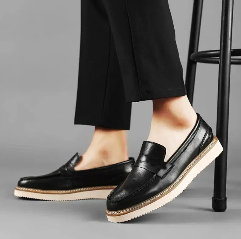 A person wearing black penny loafers with light beige soles and black pants stands next to a black stool on a gray background, showcasing a refined business casual look.