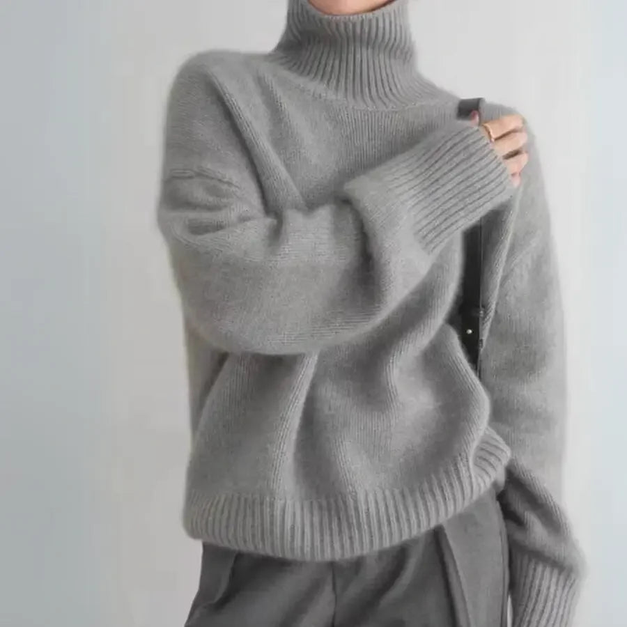 Person wearing a loose, light gray cashmere turtleneck sweater with ribbed cuffs and hem, paired with high-waisted gray pants, holding a black strap over the shoulder; head is not visible.