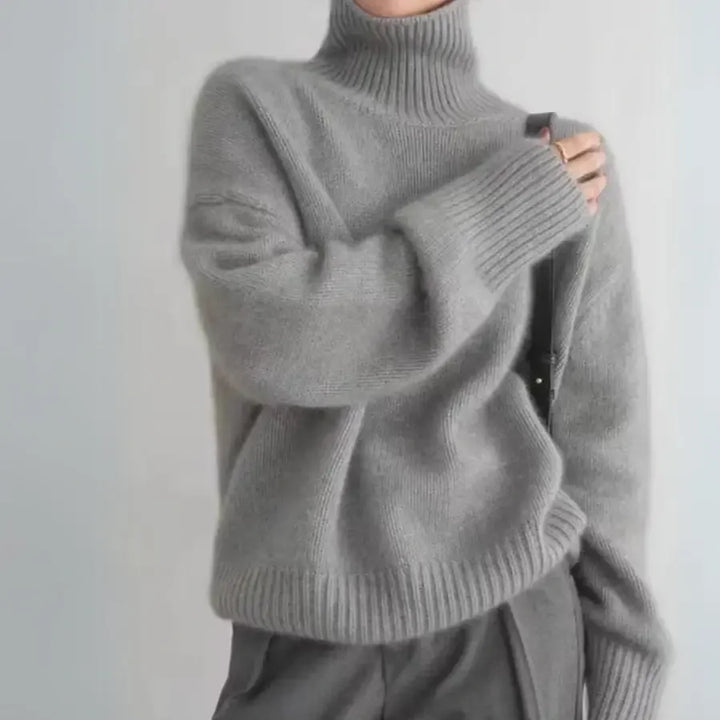Person wearing a loose, light gray cashmere turtleneck sweater with ribbed cuffs and hem, paired with high-waisted gray pants, holding a black strap over the shoulder; head is not visible.