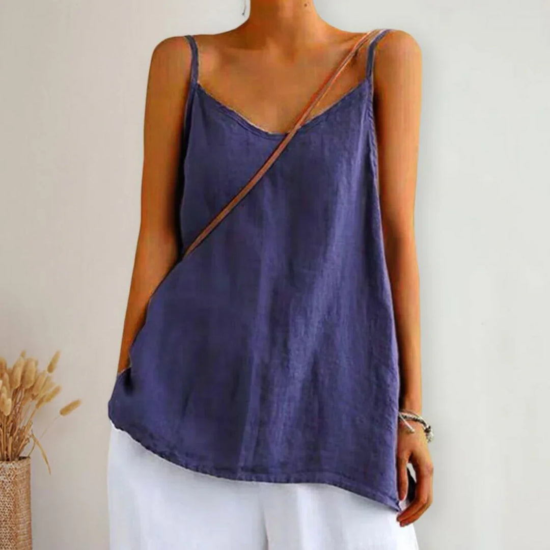 A person wears a loose-fitting, sleeveless urban-chic camisole top with thin straps and white pants, standing against a light background with a decorative vase of dried plants on the side.
