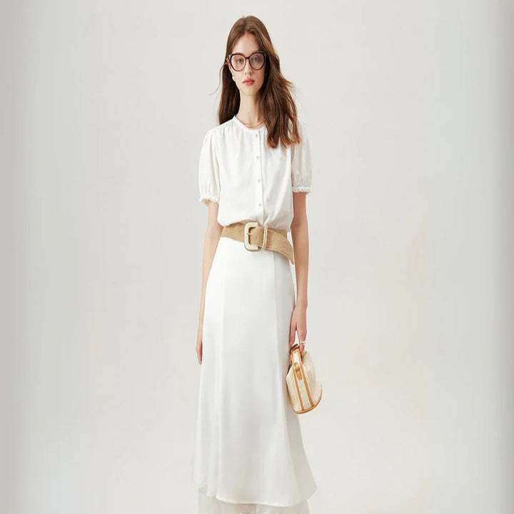 A woman wearing a white short-sleeve dress with a beige belt, holding a small round handbag. Her look is effortlessly chic—perfect for pairing with a versatile blouse or button-up blouse for added style options.