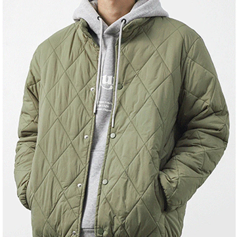 A person wearing a light olive padded jacket with a diamond-stitched pattern over a light gray hooded sweatshirt with white drawstrings, standing with hands in pockets against a plain background.