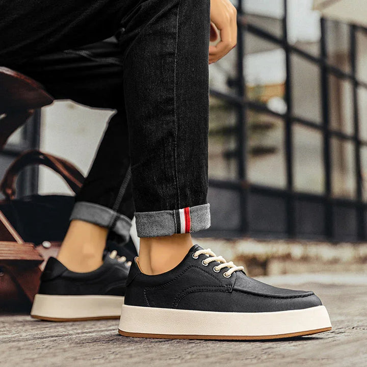 A person wearing black breathable walking shoes with white soles and laces sits outdoors on stone steps. They are dressed in black jeans with rolled-up cuffs showing a red stripe detail. A brown bag is partially visible nearby.