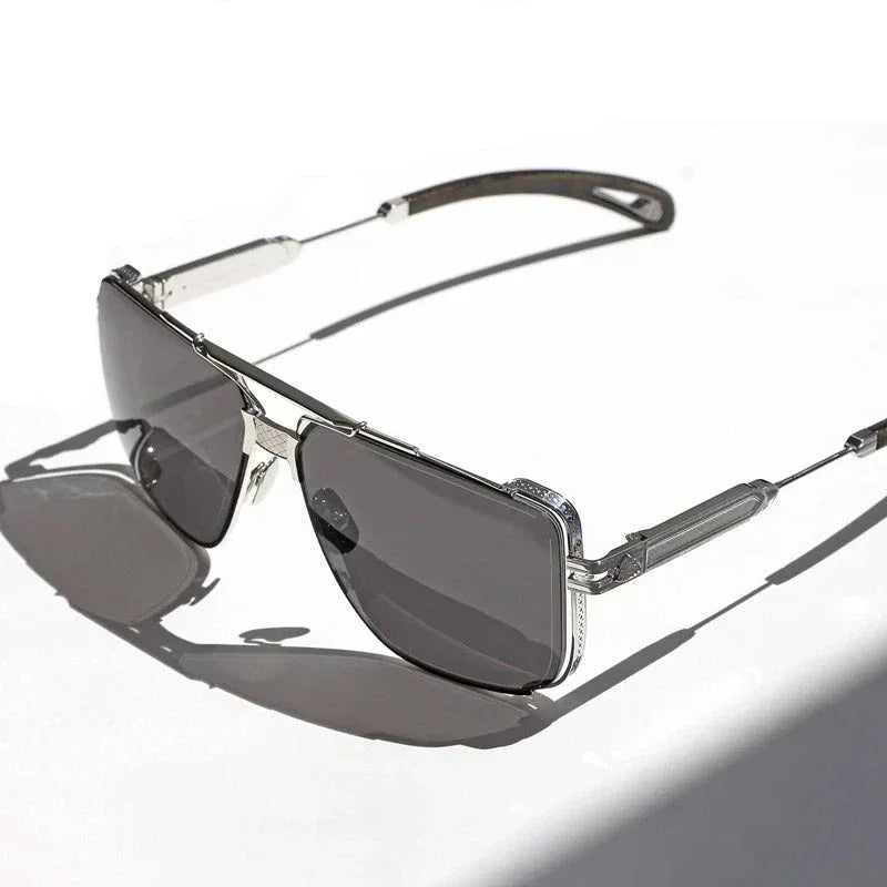 Silver-framed vintage sunglasses with dark tinted, anti-glare lenses rest on a white surface, casting a shadow to the left. The design features intricate detailing on the temples and around the lenses for added style.
