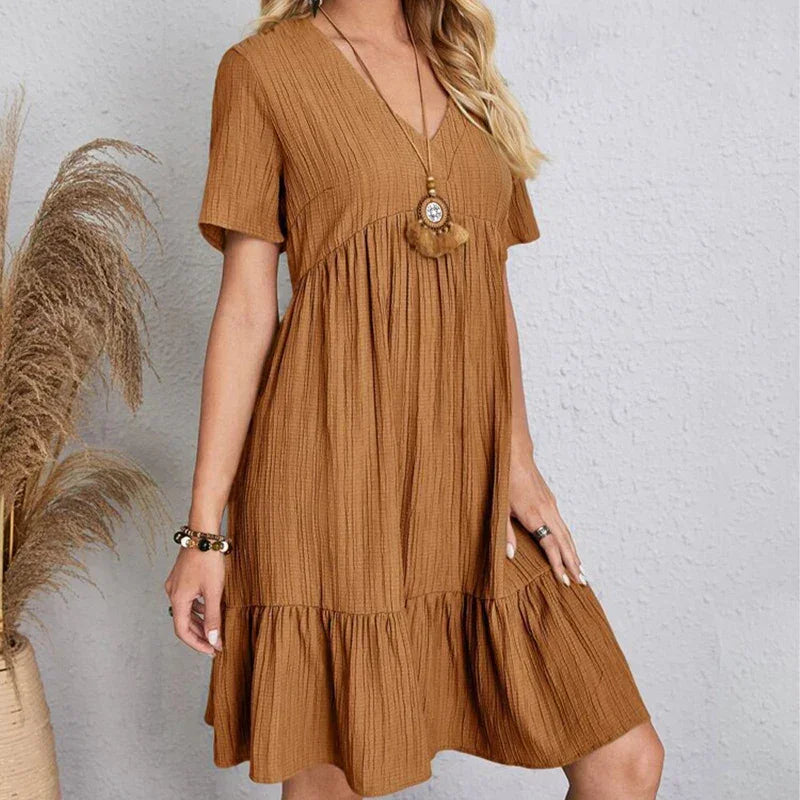 A woman wearing a loose-fitting, short-sleeved, knee-length brown dress with a ruffled hem and V-neckline stands against a light gray textured wall. She accessorizes with layered necklaces and bracelets. Pampas grass is visible to the left.