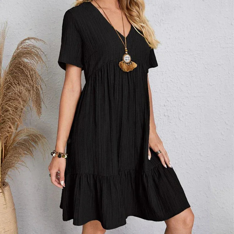 A woman in a short-sleeved, knee-length black dress with a V-neckline stands against a light textured wall. She accessorizes with a long pendant necklace and bracelet, while soft pampas grass adds a touch of nature on the left.