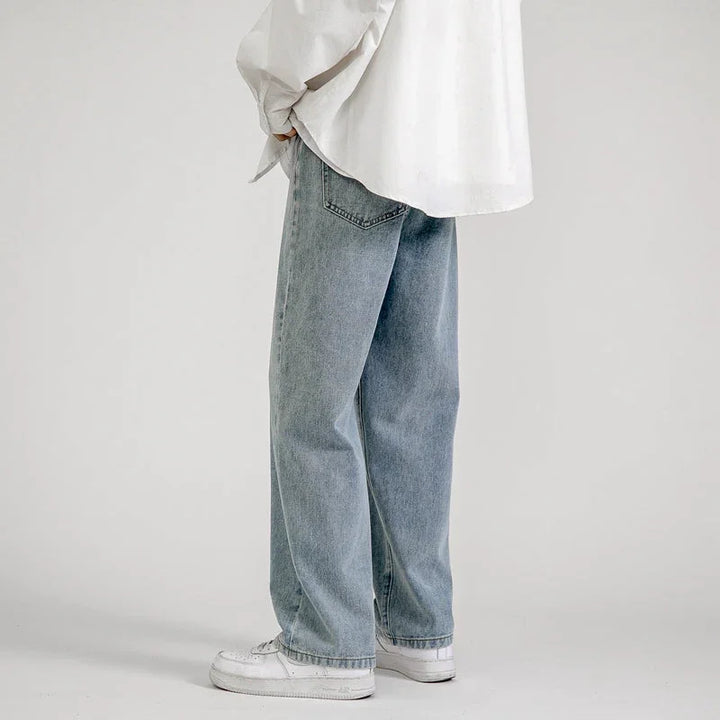 A person wearing a loose white shirt, men’s baggy jeans in a wide-leg design, and white sneakers stands against a plain light background, facing slightly away with hands in pockets for a relaxed Korean fashion vibe.