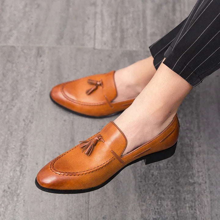 A person wearing genuine leather mens loafers with a sheepskin lining and black soles, paired with black pinstripe pants, stands on a gray tiled floor.