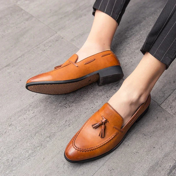 A person wearing mens loafers crafted from genuine leather with tassels and black soles, seated on a gray tiled floor, dressed in dark pinstripe pants.