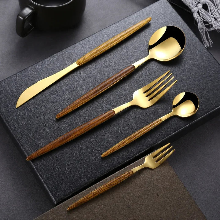 A gold plated flatware set with brown wooden handles, including a knife, tablespoon, fork, teaspoon, and dessert fork, is neatly arranged on a black textured surface with a cup and napkin in the background.