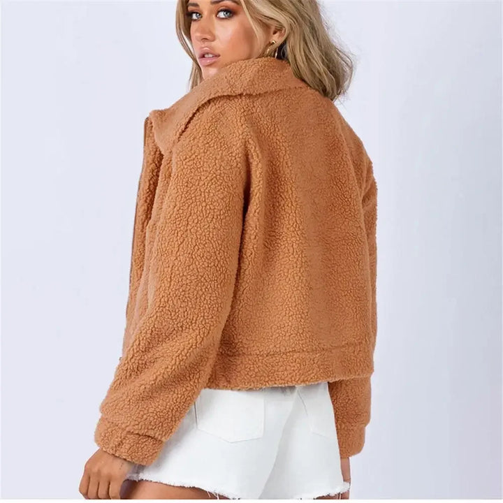 A woman wearing a tan, cropped sherpa fur jacket and white denim shorts stands facing away, looking over her shoulder against a plain light background.