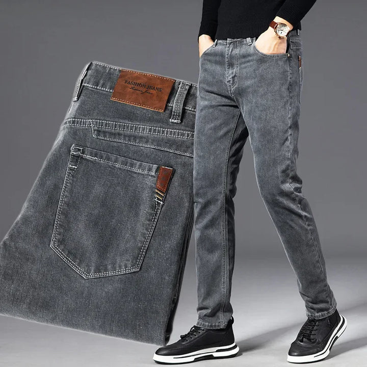 A person wearing slim-fit grey heavyweight denim jeans with hands in pockets, black sweater, watch, and black sneakers; a close-up of the jeans, showing the back pocket and leather label, is displayed beside them.