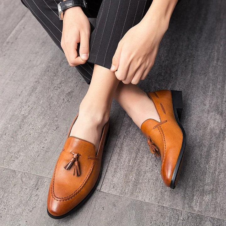 A person wearing pinstriped pants and a watch sits on a gray floor, showing off a pair of men’s loafers crafted from genuine leather with tassels.