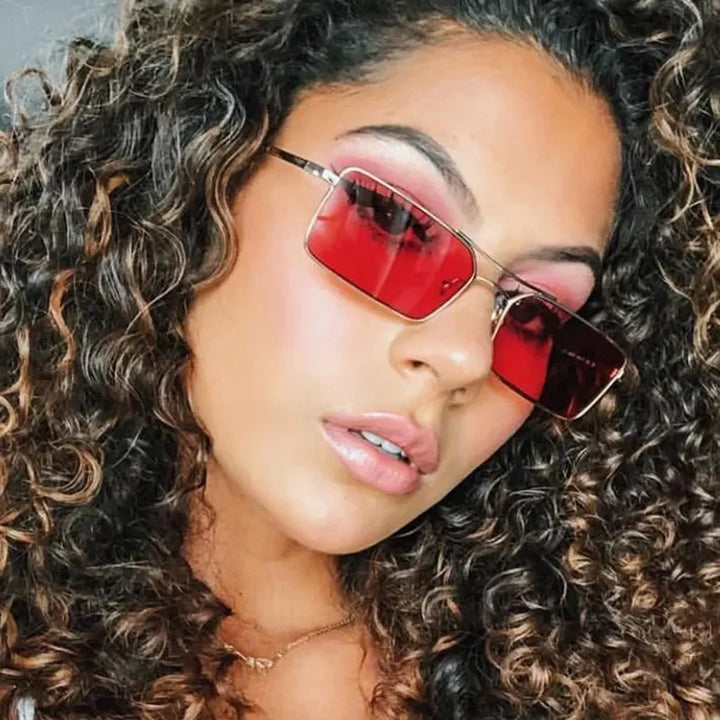 A woman with curly brown hair wears red-tinted rectangular sunglasses featuring UV400 protection and a gold necklace, tilting her head slightly. Her makeup is natural with a hint of blush, and she looks confidently at the camera.