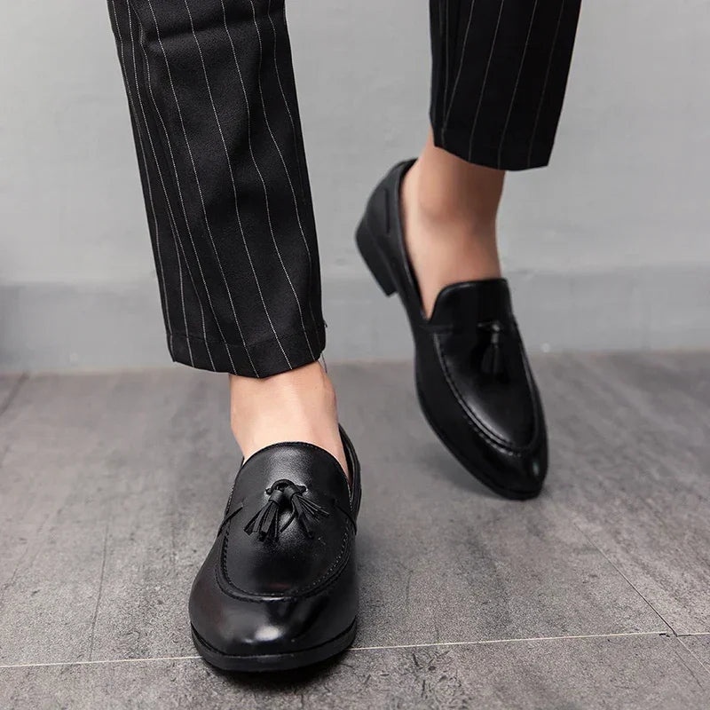 A person wearing black pinstriped pants and mens loafers with tassels stands on a gray tiled floor, showcasing genuine leather style.