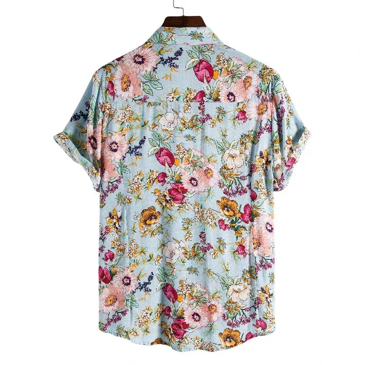A relaxed fit floral shirt with short sleeves, crafted from easy-care material, features a light blue background and a vibrant floral pattern in pink, yellow, purple, and green. Displayed on a hanger and shown from the back.