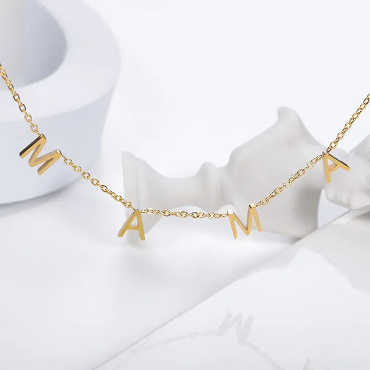 A gold minimalist jewelry piece, this stainless steel necklace features four dangling letter charms spelling MAMA, displayed against a white background with abstract white shapes.
