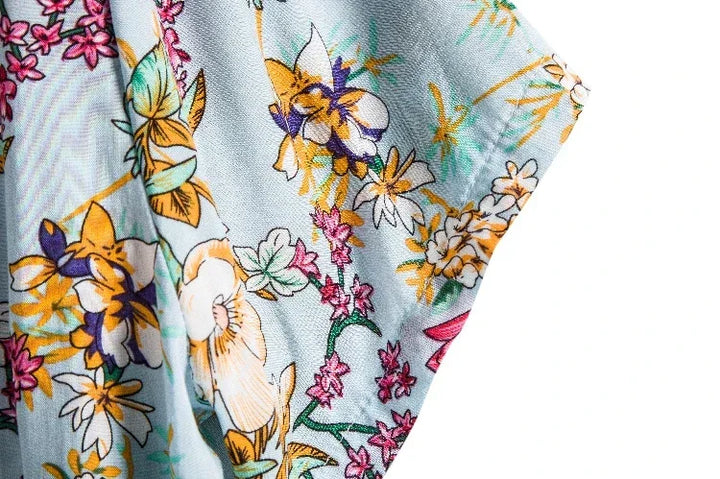 A close-up of light blue easy-care material with a colorful floral print featuring yellow, orange, pink, and purple flowers with green stems and leaves, set against a white background—perfect for a relaxed fit floral shirt.