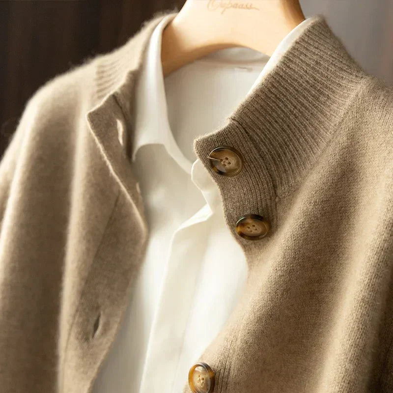 A close-up of a beige cashmere cardigan with large brown buttons, crafted from ultra soft 100% wool cashmere, worn over a white collared shirt on a hanger. The textures of the fabrics are clearly visible.