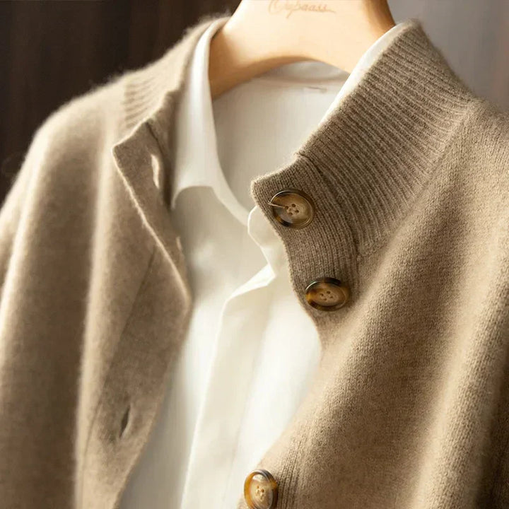 A close-up of a beige cashmere cardigan with large brown buttons, crafted from ultra soft 100% wool cashmere, worn over a white collared shirt on a hanger. The textures of the fabrics are clearly visible.