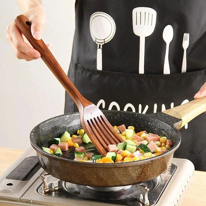 Person cooking vegetables and ham in a frying pan with a wooden spatula, wearing a black cooking apron, Rovelli kitchenware.
