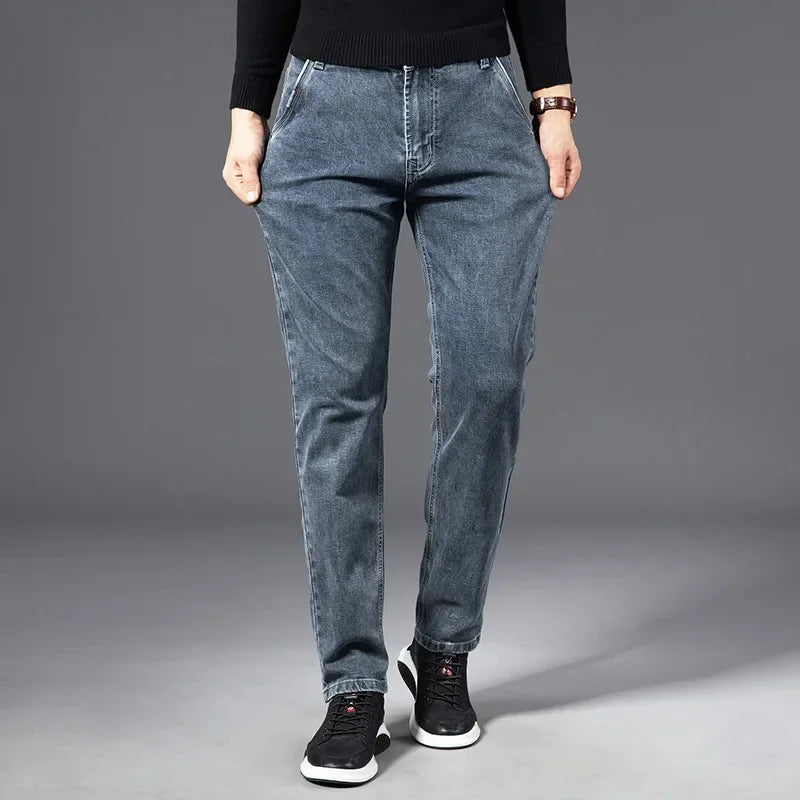A person wearing a black sweater, straight-leg blue jeans, and black sneakers stands with hands in pockets against a plain gray background.